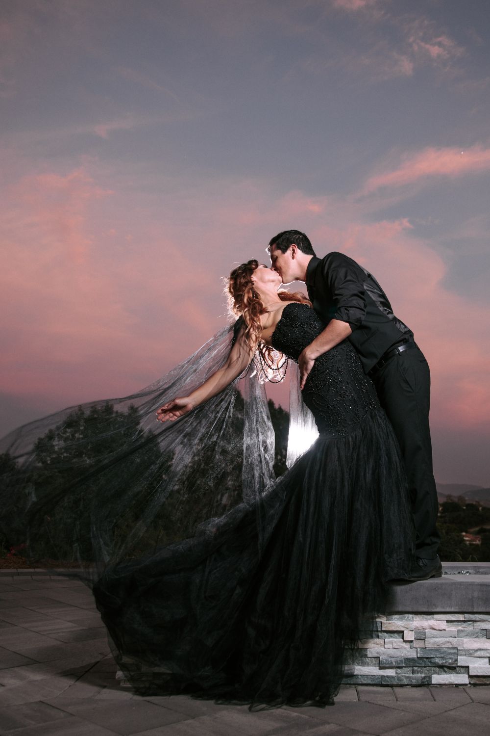 Bride wearing black wedding dress kissing groom in front of California sunset.